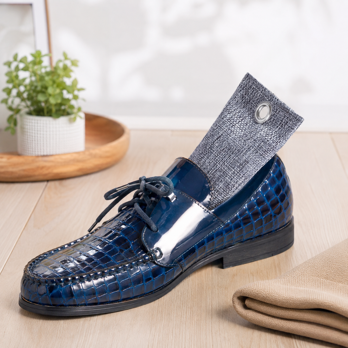 A navy blue crocodile-embossed leather loafer with a matching dark gray textured sock tucked into the open back, styled on a light wood surface with a small potted plant and beige fabric in the background.